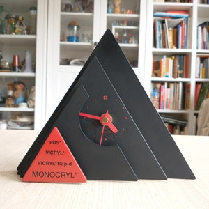 May include: A black triangular clock with a red face and hands. The clock is made of three layers of black triangles. The bottom layer has a red triangle with the words "PDS", "VICRYL", "VICRYL Rapid", and "MONOCRYL" printed on it.