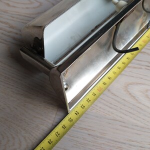 May include: A silver metal wall light fixture with a white glass shade. The fixture is mounted on a wall and has a tape measure next to it.