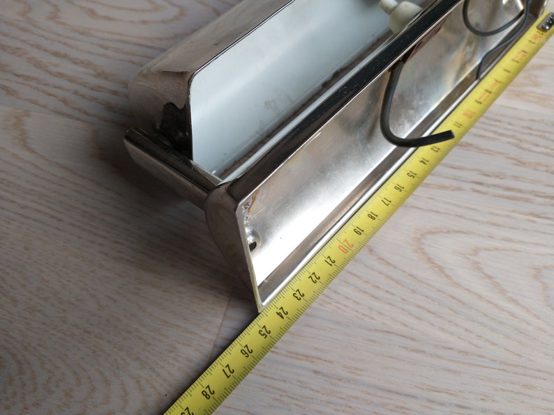 May include: A silver metal wall light fixture with a white glass shade. The fixture is mounted on a wall and has a tape measure next to it.