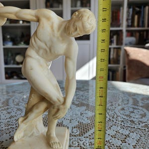 May include: A cream-colored marble-style statue of a discus thrower in a dynamic athletic pose, standing on a square base. A yellow measuring tape is positioned vertically next to the figure, indicating a height of approximately 7.75 inches.