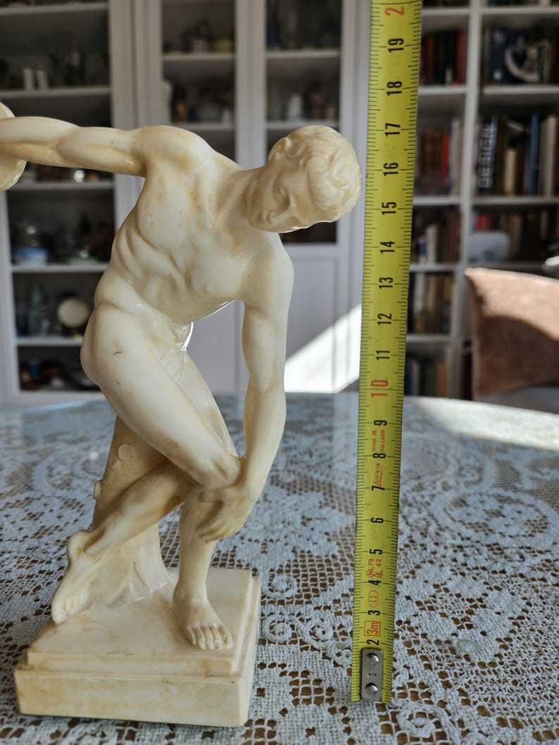 May include: A cream-colored marble-style statue of a discus thrower in a dynamic athletic pose, standing on a square base. A yellow measuring tape is positioned vertically next to the figure, indicating a height of approximately 7.75 inches.
