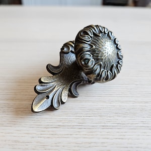 Könnte beinhalten: Ein dekorativer Haken aus Metall mit einem verzierten Design. Der Haken ist in Bronze gehalten und hat einen runden Knopf mit einem floralen Muster.