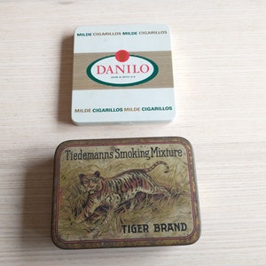 Puede incluir: Dos latas de tabaco vintage. La primera lata es blanca con detalles rojos y verdes y dice "Danilo" en negro. La segunda lata es marrón con una ilustración de tigre y dice "Tiedemanns Smoking Mixture Tiger Brand".