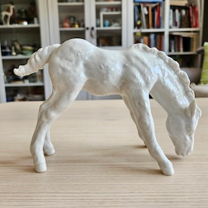 May include: A white porcelain figurine of a horse foal with its head down, as if grazing. The figurine is detailed with textured fur and a glossy finish.