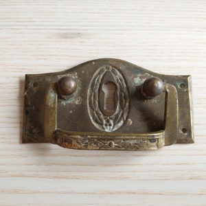 Puede incluir: Tirador de cajón de latón antiguo con una cerradura y un diseño ornamentado. El tirador tiene una parte superior curva y una manija rectangular.