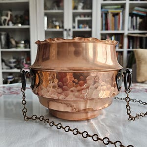 May include: A copper hanging planter with a hammered finish and two black metal hooks. The planter is suspended by a chain.
