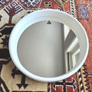 Op de afbeelding: Een ronde spiegel met een witte lijst en een reflecterend oppervlak. De spiegel heeft een eenvoudig ontwerp en een klein, goudkleurig embleem aan de bovenkant. Hij hangt aan een dunne, goudkleurige draad.