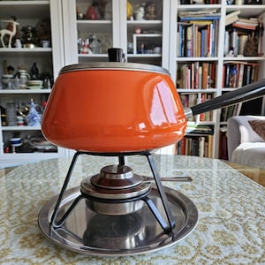 May include: An orange fondue pot with a black handle and lid, set on a metal stand with a burner. The pot is round with a shiny finish. The stand sits on a silver tray. A classic piece for entertaining.