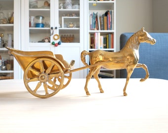 May include: A gold-colored brass horse-drawn carriage figurine. The carriage has a single wheel and is pulled by a horse with its head turned to the side. The figurine is on a white surface.