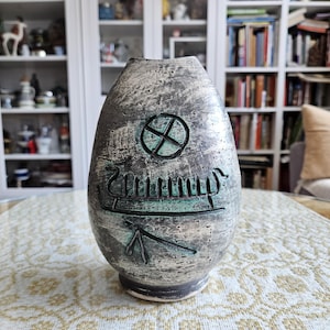 Peut inclure: Un vase en céramique texturé, gris et sarcelle, avec un design unique. Le vase présente une croix sarcelle dans un cercle, un navire viking sarcelle et d'autres symboles sarcelle. Le vase est sur une table avec une nappe à motifs.