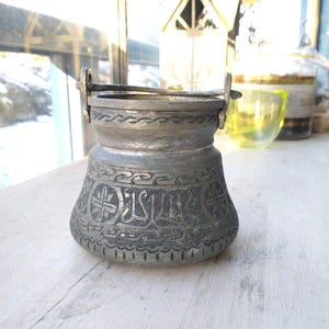Puede incluir: Una olla de metal plateado con un diseño adornado y un asa. La olla está sobre una superficie de madera.