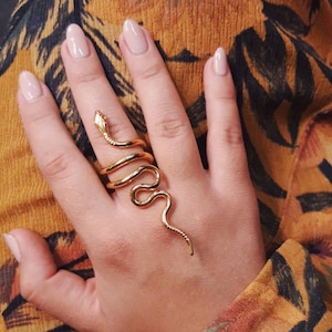 May include: A gold snake ring with a coiled body and a pointed tail. The ring is worn on a finger with nude nail polish.