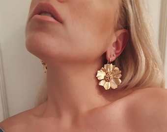May include: A pair of gold flower-shaped earrings with a textured finish. The earrings are dangling and have a small gold bead in the center of each flower.