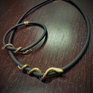 May include: A black rubber necklace with a gold snake charm. The necklace is on a dark wooden surface.