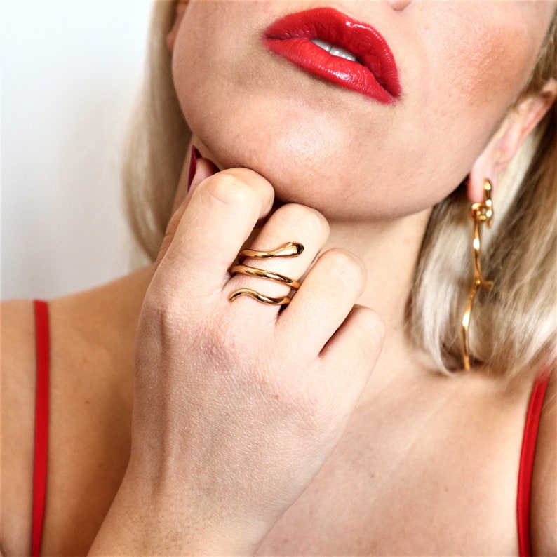 May include: A gold snake ring with a coiled design. The ring is worn on the index finger of a hand.