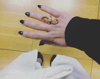 May include: A hand with black nail polish is wearing a gold ring with a unique design. The ring is on the middle finger. The hand is wearing a black long-sleeved shirt.