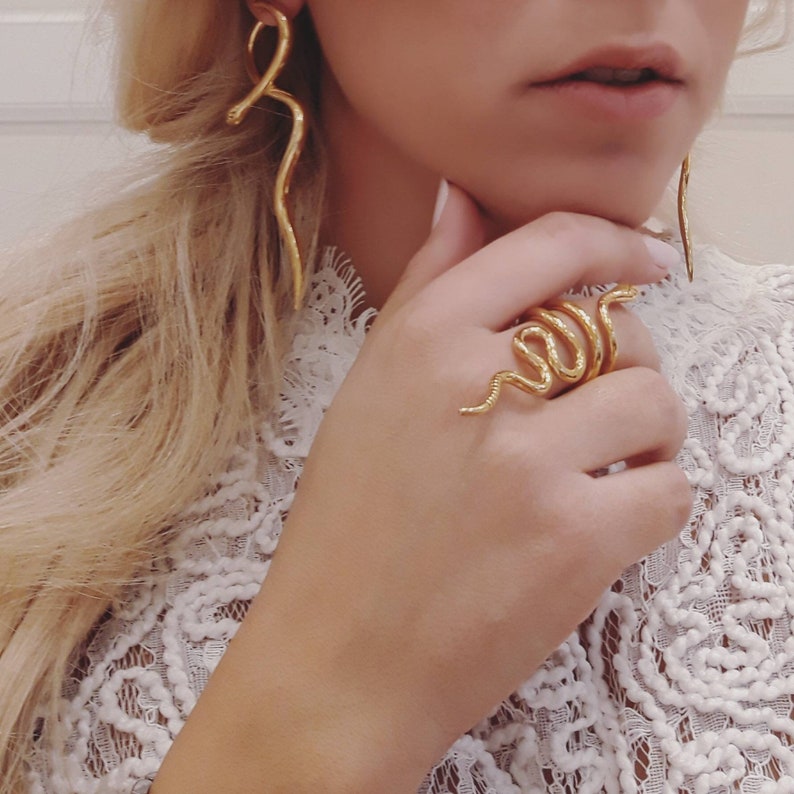 May include: A gold snake ring with a textured finish. The ring is worn on the index finger of a hand.