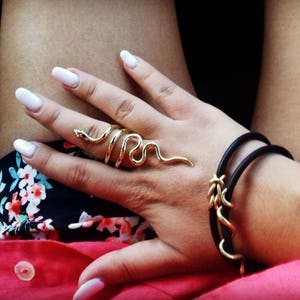 May include: A gold snake ring with a black leather bracelet on a person's wrist. The person is wearing a floral print shirt and a red shirt.