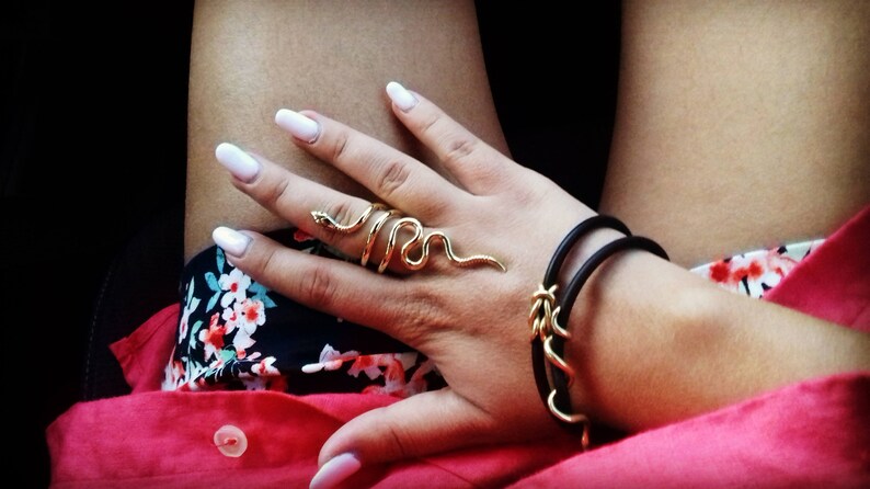 May include: A gold snake ring with a black leather bracelet on a person's wrist. The person is wearing a floral print shirt and a red shirt.