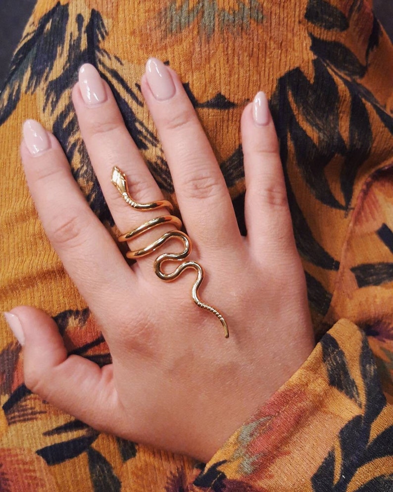 May include: A gold snake ring with a coiled body and a pointed tail. The ring is worn on a finger with nude nail polish.