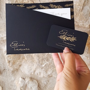 May include: Black gift envelope with gold leaf accents and a handwritten "Thank you" note. A black gift card with gold lettering reads "Ellina's Treasures" with a gold olive branch design.