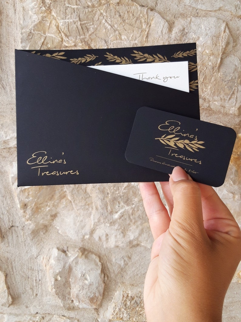 May include: Black gift envelope with gold leaf accents and a handwritten "Thank you" note. A black gift card with gold lettering reads "Ellina's Treasures" with a gold olive branch design.
