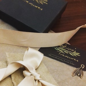 May include: A small, brown, rectangular box with a gold leaf design and the text "Elina's Treasures" on the top. The box is tied with a cream-colored ribbon and is sitting on a brown surface.
