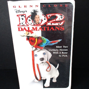 May include: A VHS tape for the movie 102 Dalmatians, featuring Glenn Close as Cruella de Vil. The cover shows a white puppy with a red leash and a red, green, and blue parrot.
