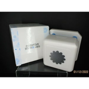 Puede incluir: Un troquel de papel blanco con forma de flor de Creative Memories con un diseño de flor gris. El troquel está junto a una caja blanca con las palabras "Creative Memories" impresas. La caja tiene un estampado floral.