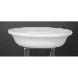May include: A white, oval ceramic bowl with a rounded rim. The bowl is set against a dark background, and appears to be sitting on a clear stand. The bowl's smooth surface reflects light, highlighting its clean design.
