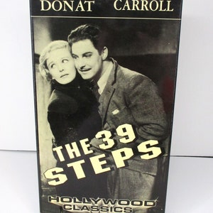May include: A black and white photograph of Robert Donat and Madeleine Carroll on the front of a DVD case for the film "The 39 Steps". The DVD case is black with gold lettering and the title "Hollywood Classics Collectors Edition".