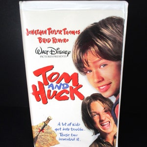 Puede incluir: Una cinta VHS de la película "Tom y Huck" de Disney, con Jonathan Taylor Thomas y Brad Renfro. La carátula blanca presenta el título en rojo y azul, y fotos de los actores.
