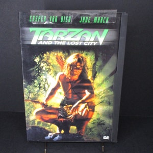Può includere: Custodia DVD per il film Tarzan e la città perduta, con uno sfondo di giungla verde e un uomo muscoloso con un perizoma che tiene una lancia. Il titolo è in grandi lettere verdi e in grassetto. Gli attori Casper Van Dien e Jane March sono elencati nella parte superiore della custodia.