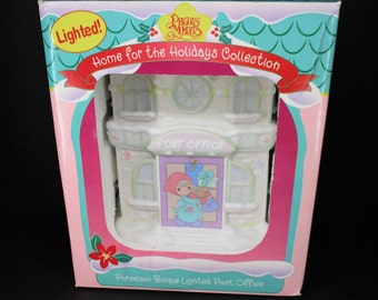May include: A porcelain bisque lighted post office figurine from the Precious Moments Home for the Holidays Collection. The figurine features a small girl in a red hat and blue coat carrying a bag of gifts. The post office has a sign that reads "Post Office".