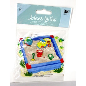 Puede incluir: Un paquete de pegatinas dimensionales de Jolee's by You. Las pegatinas presentan una escena de arenero con un arenero azul, un camión de juguete, un cubo, una pala y otros accesorios. El paquete incluye el texto "ETIQUETAS DIMENSIONALES".