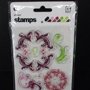 May include: A set of six clear stamps with floral designs in shades of pink, purple, and green. The stamps are packaged in a clear plastic bag with the text "whisper stamps" and "BASICGREY" printed on the front.
