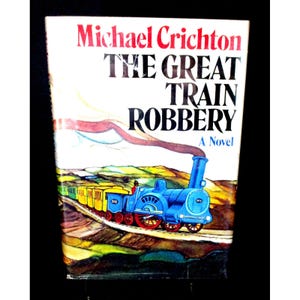 May include: A vintage paperback book titled "The Great Train Robbery" by Michael Crichton. The cover shows a blue steam train with yellow carriages on a track, set against a landscape of green hills and a white sky. The title is in large black letters.