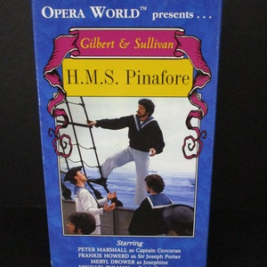 May include: A vintage VHS tape for the Gilbert & Sullivan opera "H.M.S. Pinafore." The blue box features a colorful illustration of sailors on a ship, with the title in large yellow letters. The tape is from Opera World.