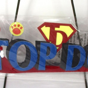 Może przedstawiać: Zestaw 5 naklejek trójwymiarowych z czerwono-niebiesko-czarną wstęgą "Top Dog" z czerwono-żółtym logo Supermana. Naklejki zawierają również odciski łap w kolorze czerwonym, żółtym i niebieskim.