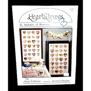 May include: A white paper with the title "HeartStrings: An Alphabet of Memories" is displayed. The image features framed heart-shaped cross-stitch samplers, a heart garland, and decorative pillows. The Artists Collection presents Pat Thode, Designer.