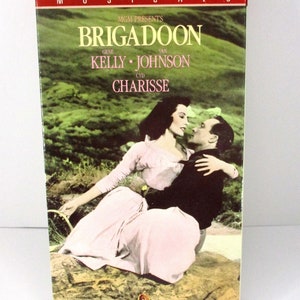 Könnte beinhalten: Ein VHS-Kassetten-Etui für den Musikfilm "Brigadoon" mit Gene Kelly und Van Johnson. Das Cover zeigt ein Foto von Kelly und Cyd Charisse, die sich in einem Feld umarmen.