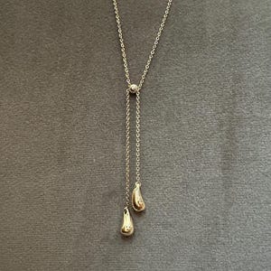 May include: A delicate gold-colored necklace with a Y-shaped design. The necklace features a small, round accent at the center and two teardrop-shaped pendants at the end of the chain. The necklace is displayed against a gray background.