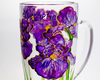 May include: A glass mug with a hand-painted design of purple iris flowers with gold accents. The mug has the name "Lecretia" written on it.