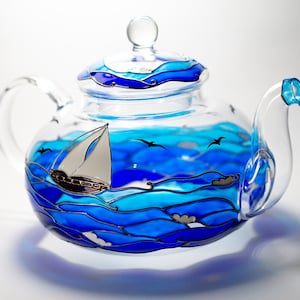 Puede incluir: Una tetera de vidrio con un diseño pintado en azul y blanco de un velero en el océano. La tetera tiene una tapa de vidrio transparente y un asa.