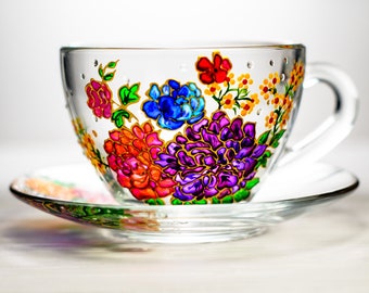 May include: A glass teacup and saucer with a hand-painted floral design. The cup features a colorful bouquet of flowers in shades of pink, purple, blue, yellow, and green. The saucer has a matching floral design.