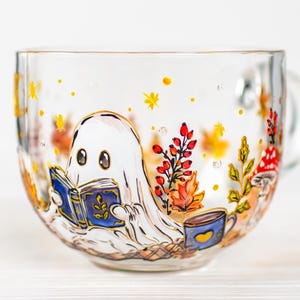 May include: A clear glass mug featuring a hand-painted illustration of a white ghost reading a blue book. The design includes autumn-themed elements such as red berries, orange leaves, a small blue mug with a yellow heart, and a red-spotted mushroom.