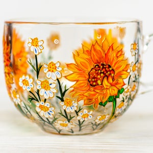 May include: A clear glass mug featuring hand-painted floral designs. The artwork displays a large, vibrant orange and yellow sunflower alongside smaller white daisies with yellow centres and green stems, wrapping around the exterior surface.