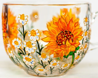May include: A clear glass teacup with a large, hand-painted sunflower and smaller white and yellow daisies. The sunflower is a vibrant orange and yellow, with a brown center. The cup has a handle and is set against a white background.