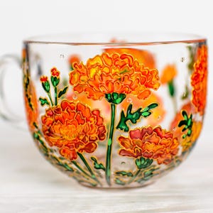 May include: A clear glass teacup with a handle, adorned with hand-painted orange and yellow marigold flowers. The floral design is outlined in gold, with green stems and leaves. The cup is set against a white background.