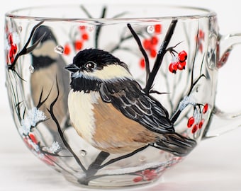 May include: A clear glass mug featuring a hand-painted design of a black-capped chickadee perched on a dark branch. The artwork includes clusters of bright red berries and white snow accents on the branches, wrapping around the exterior surface.